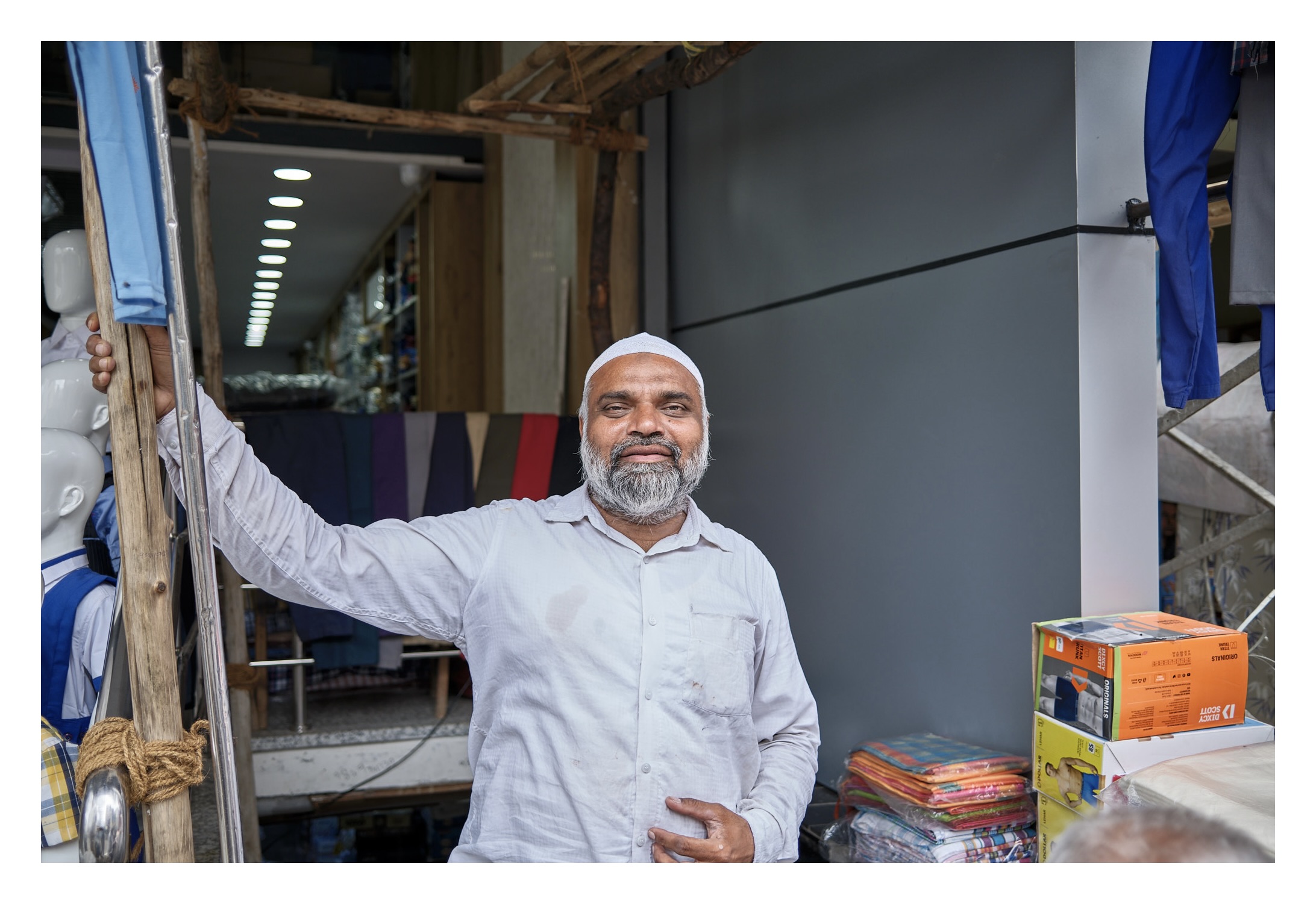 a muslim man posing in front of his store wearing a white skull cap and white dress