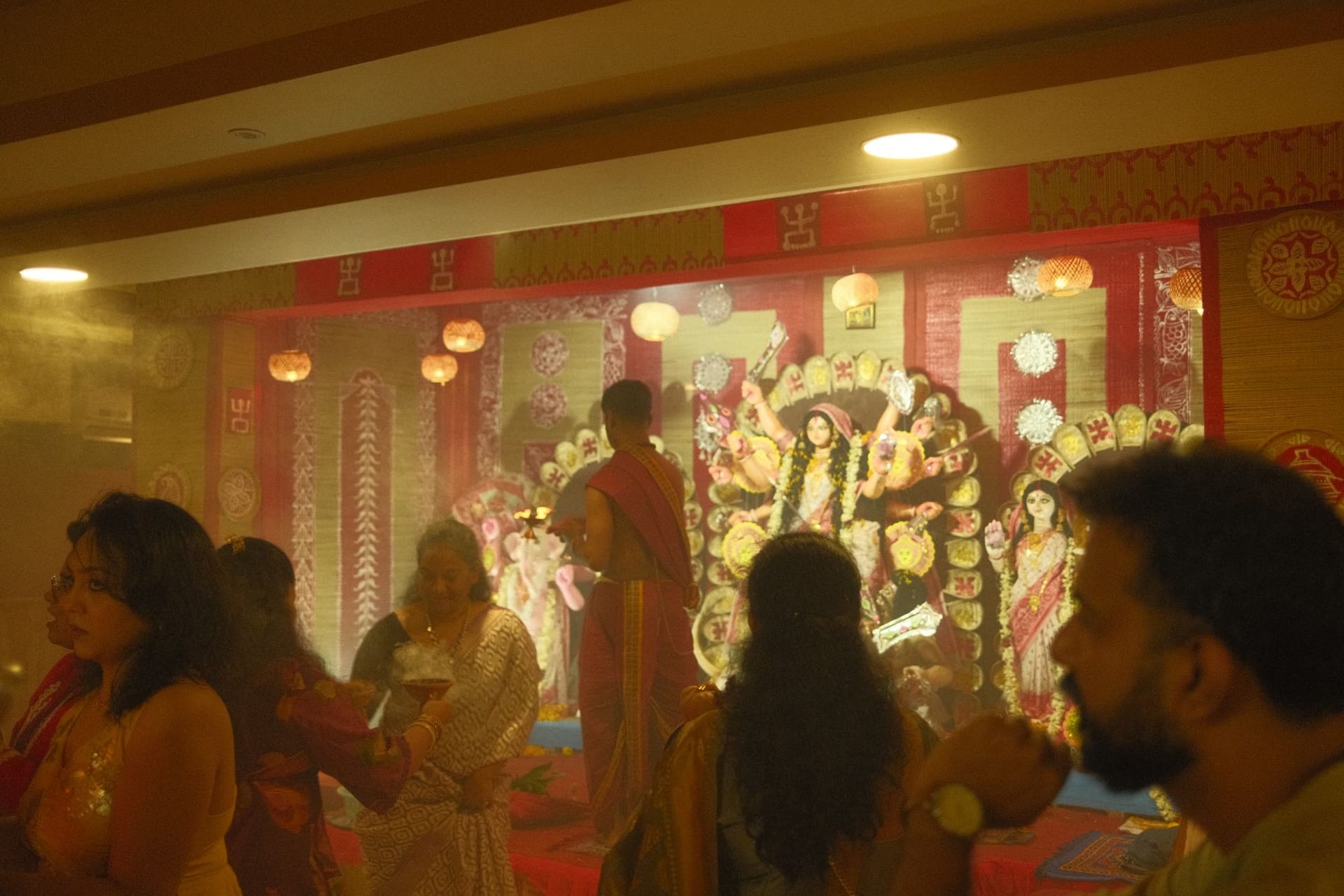 People offering anjali in front of the Durga idol, with incense smoke in the air and a priest in a red dhoti at the front