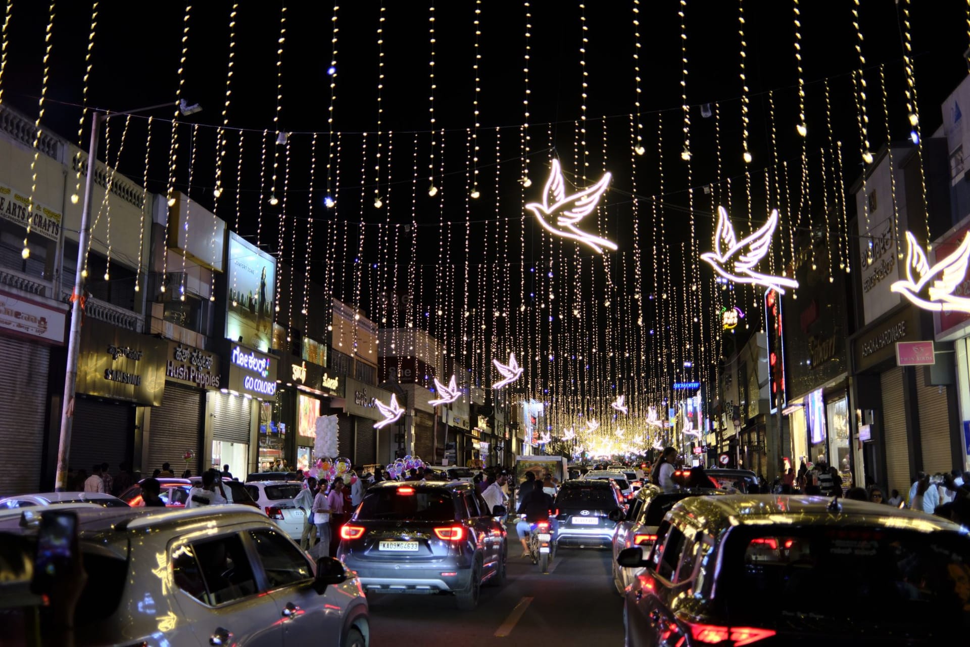 A Mysore street at night under curtains of fairy lights and glowing bird shapes, with cars and people filling the road below