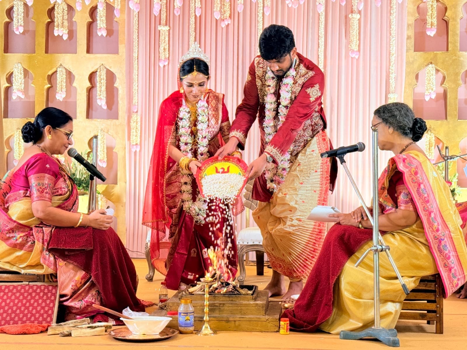 Sreejita and Rajath pouring puffed rice into the sacred fire from a painted kula, with the two priestesses seated on either side reading the mantras into microphones